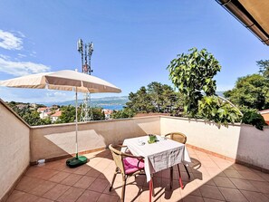 A terrace with a table and chairs, a table umbrella, a potted plant, and a view of the sea and buildings. - Apartment Dragica by Interhome (Polje)