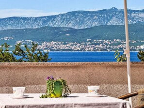 A table with a cup and saucer, a vase with flowers, and a view of a coastal town with mountains in the background. - Apartment Dragica by Interhome (Polje)