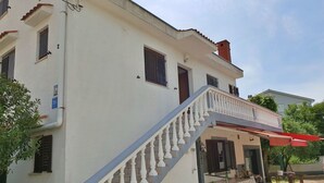 Um edifício de dois andares com fachada branca, varanda com toldo vermelho e escadaria com balaústres brancos.