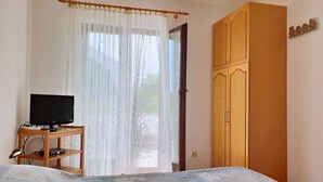 Quarto com cama, televisão sobre suporte de madeira, guarda-roupa e janela com cortinas leves.