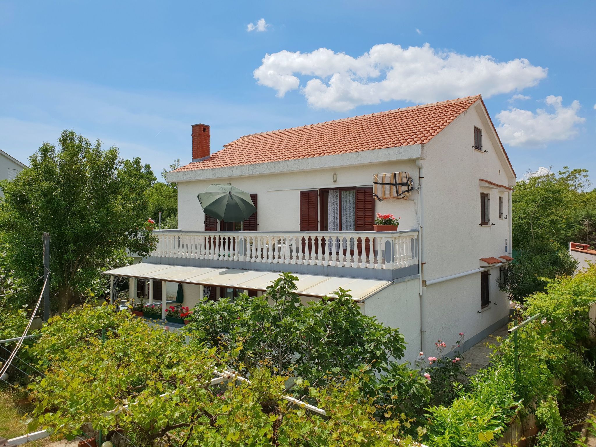 A two-story house with a red-tiled roof, a balcony with a white railing, and a garden in the foreground.