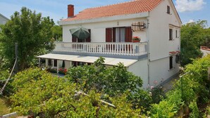 Uma casa de dois andares com telhado de telhas vermelhas, uma varanda com grades brancas e um jardim ao primeiro plano.