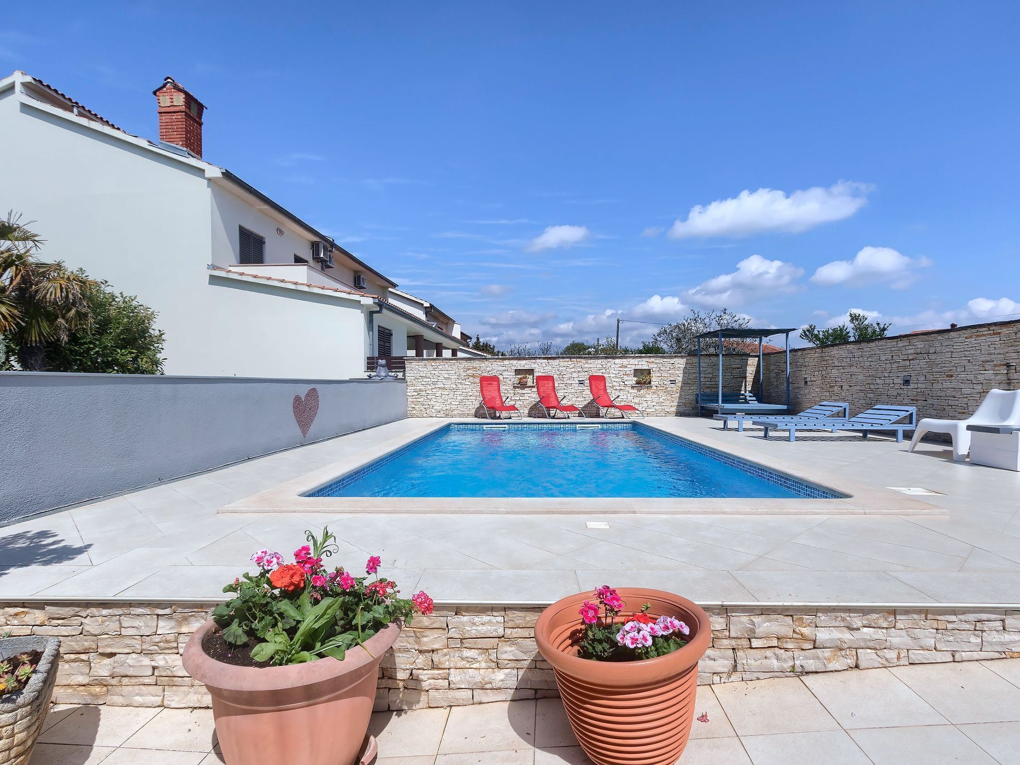 A swimming pool with red lounge chairs, a stone wall, and potted plants.