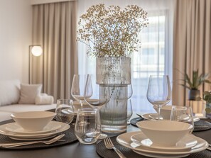 A dining table set with white plates, bowls, and wine glasses, a vase with flowers, and a modern lamp in a room with a sofa and curtains. - White Space am Meer by Interhome (Swinoujscie)
