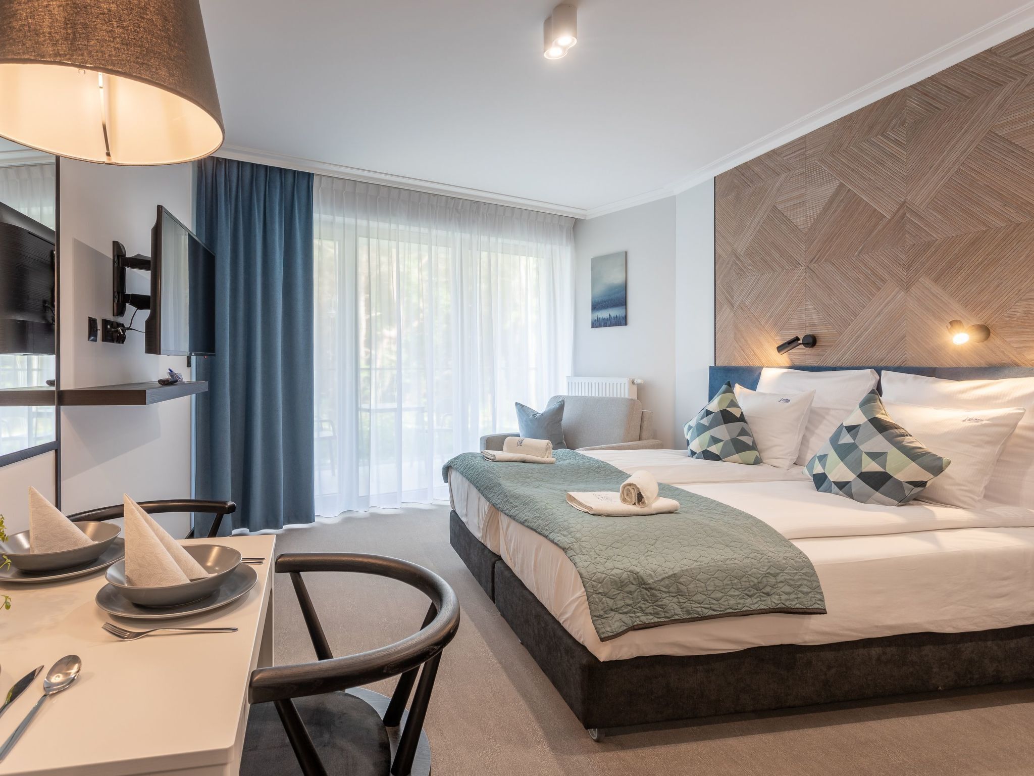 Une chambre d’hôtel moderne équipée d’un lit, de tables de chevet, d’un bureau avec une chaise, d’une télévision et d’une grande fenêtre avec des rideaux.
