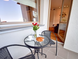 Ein kleiner runder Tisch mit Glasplatte, zwei Stühle, eine Vase mit roten Blumen, ein Croissant und eine weiße Tasse auf einem Balkon mit Blick ins Innere.