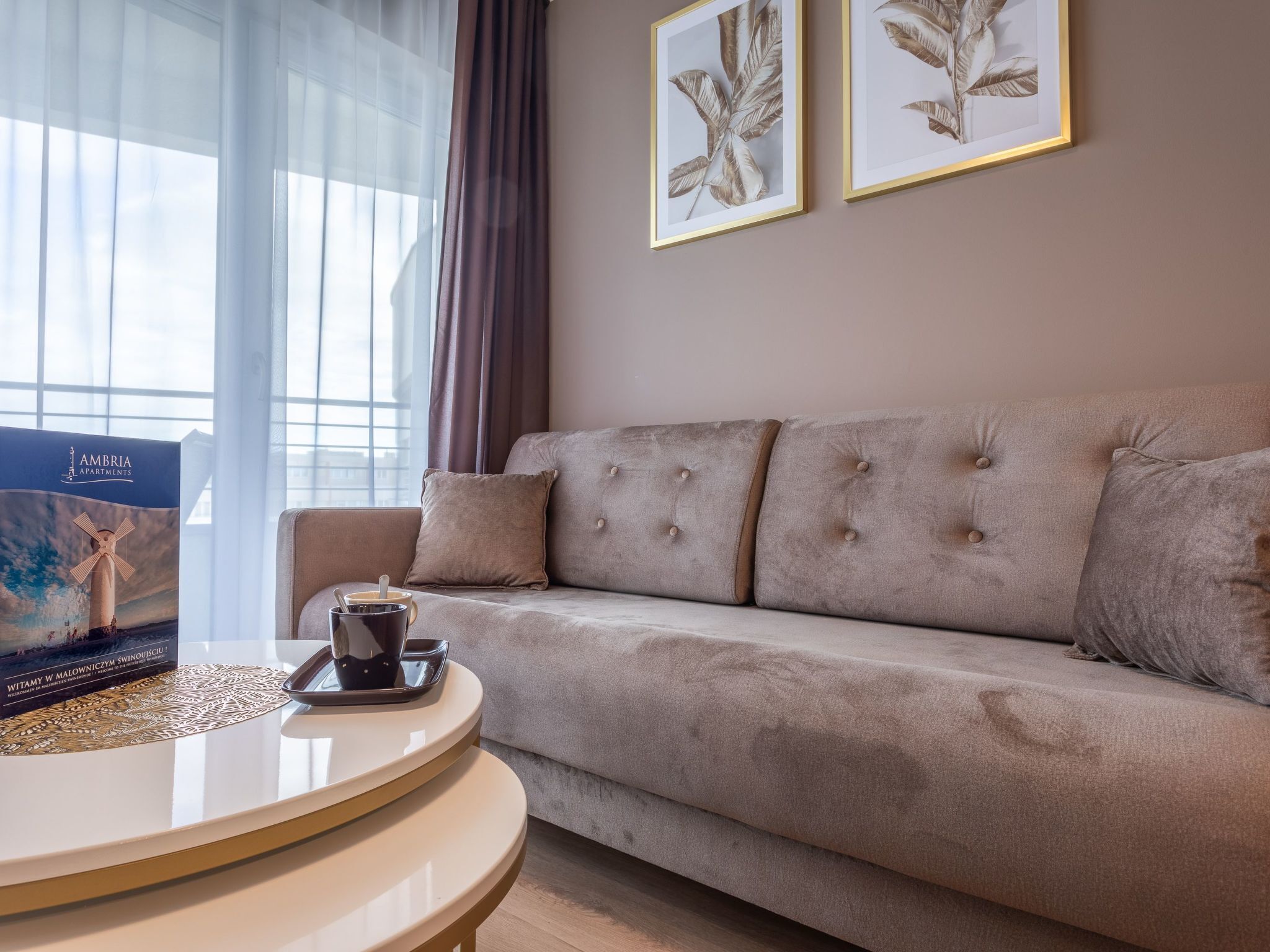 Ein Wohnzimmer mit einem beigefarbenen Sofa, einem runden Couchtisch und zwei gerahmten Blätterbildern an der Wand.