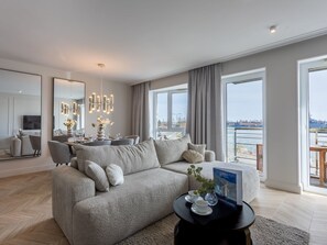 Ein modernes Wohnzimmer mit einem grauen Sofa, einem Essbereich mit einer Deckenleuchte und großen Fenstern mit Blick auf ein Gewässer.