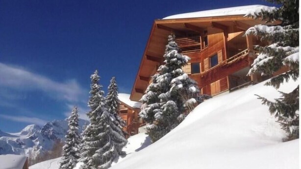 Una cabaña de madera con balcón, rodeada de árboles cubiertos de nieve y montañas al fondo.