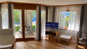 Un salón con suelo de madera, un sofá, un televisor sobre un mueble de madera y una puerta corrediza de cristal que da a un área exterior con una mesa y sillas.