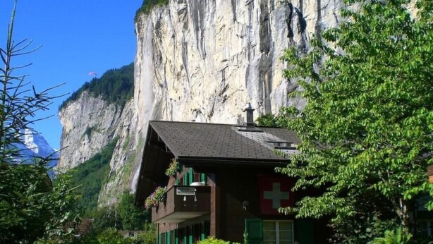 A mountain lodge with a steep roof, a Swiss flag, and a view of a rocky cliff. - Margrit West by Interhome (Lauterbrunnen)
