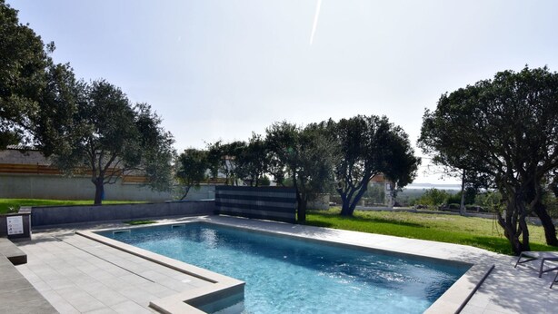 Une piscine rectangulaire, bordée d’une terrasse carrelée, entourée d’arbres et d’une pelouse.