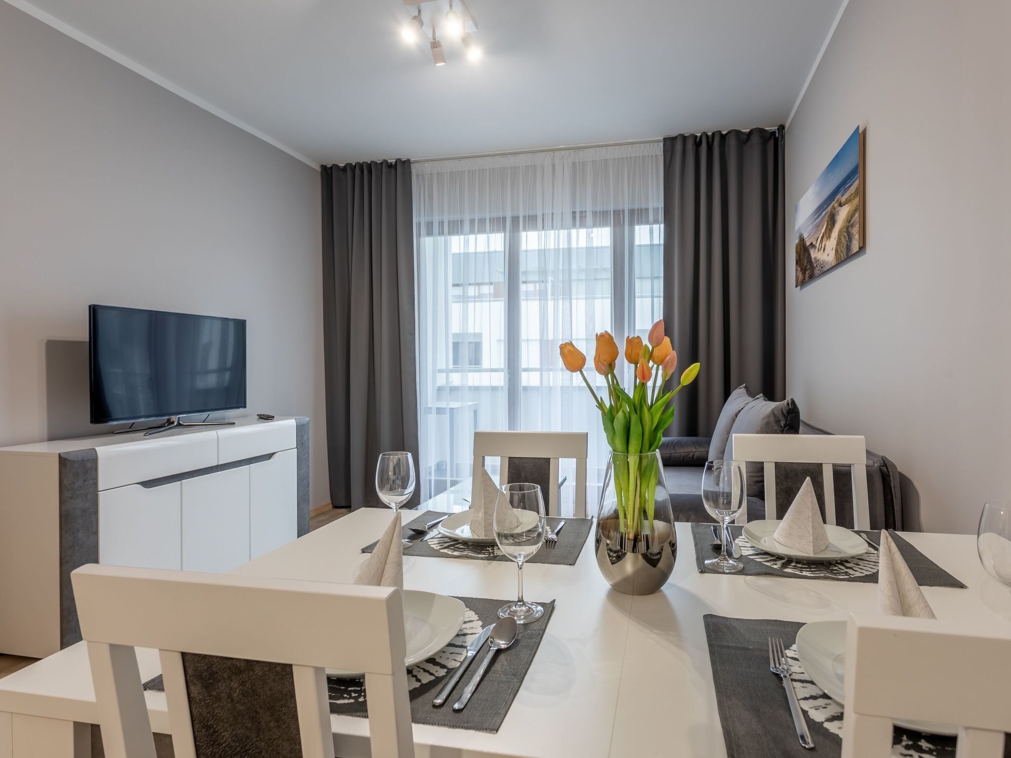 Een moderne eetruimte met een witte tafel gedekt voor vier personen, een flatscreen televisie op een witte kast en een groot raam met grijze gordijnen.