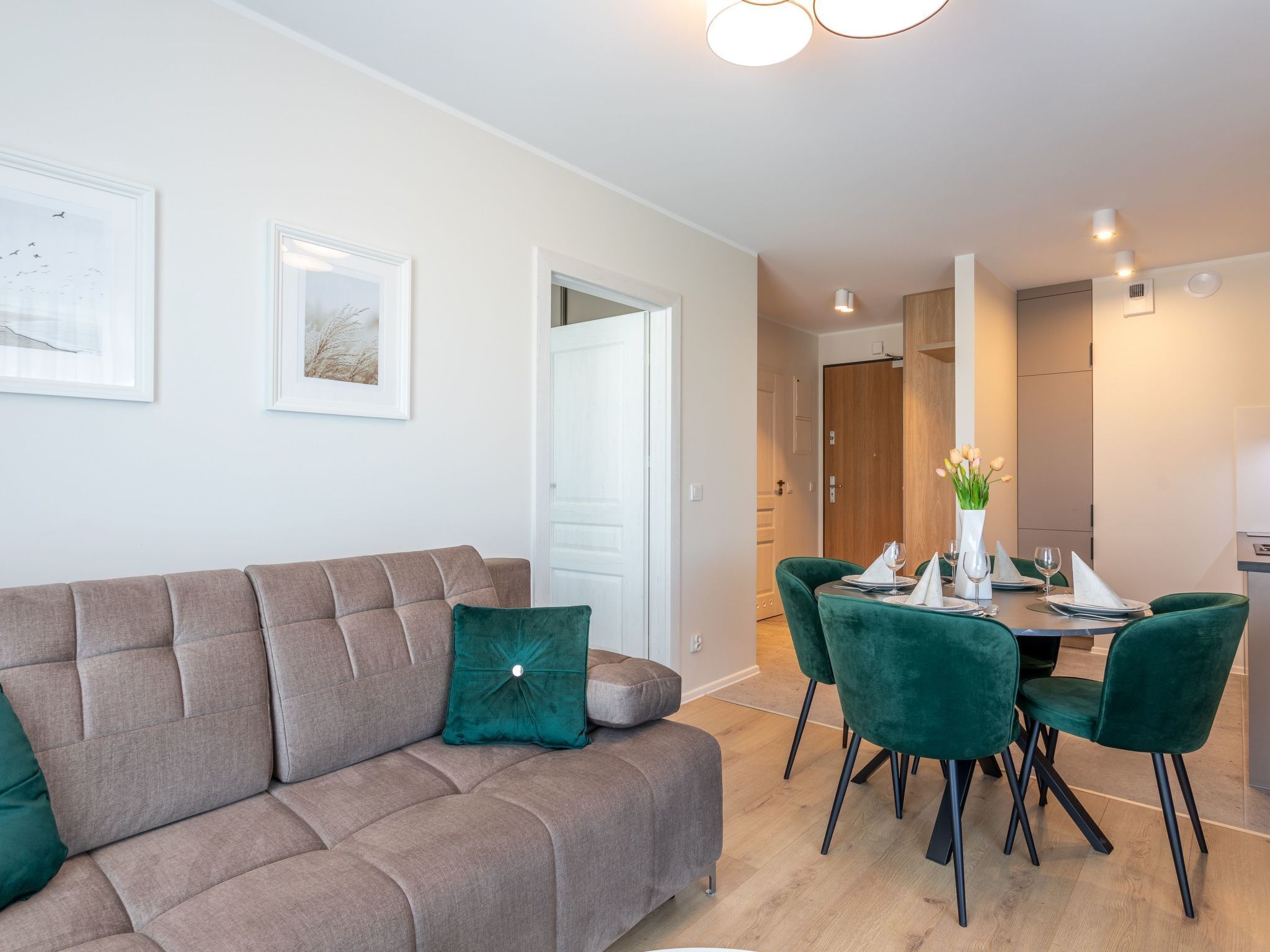 A modern living room with a beige sofa, green cushions, and a dining area with green chairs and a table set for two.