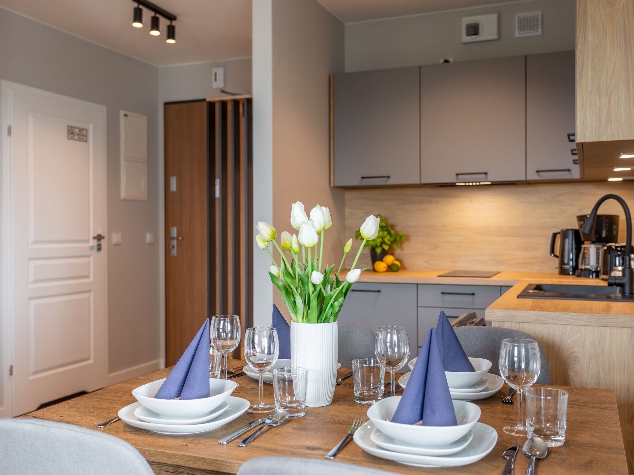 Un coin repas avec une table en bois dressée pour quatre personnes, des assiettes blanches, des serviettes bleues et des verres transparents.