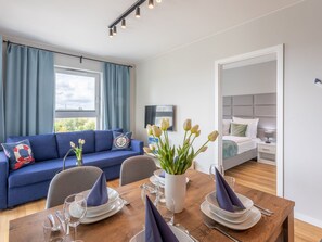 Ein modernes Wohnzimmer mit einem blauen Sofa, einem Esstisch mit Tellern und Gläsern sowie Stadtblick durch große Fenster.
