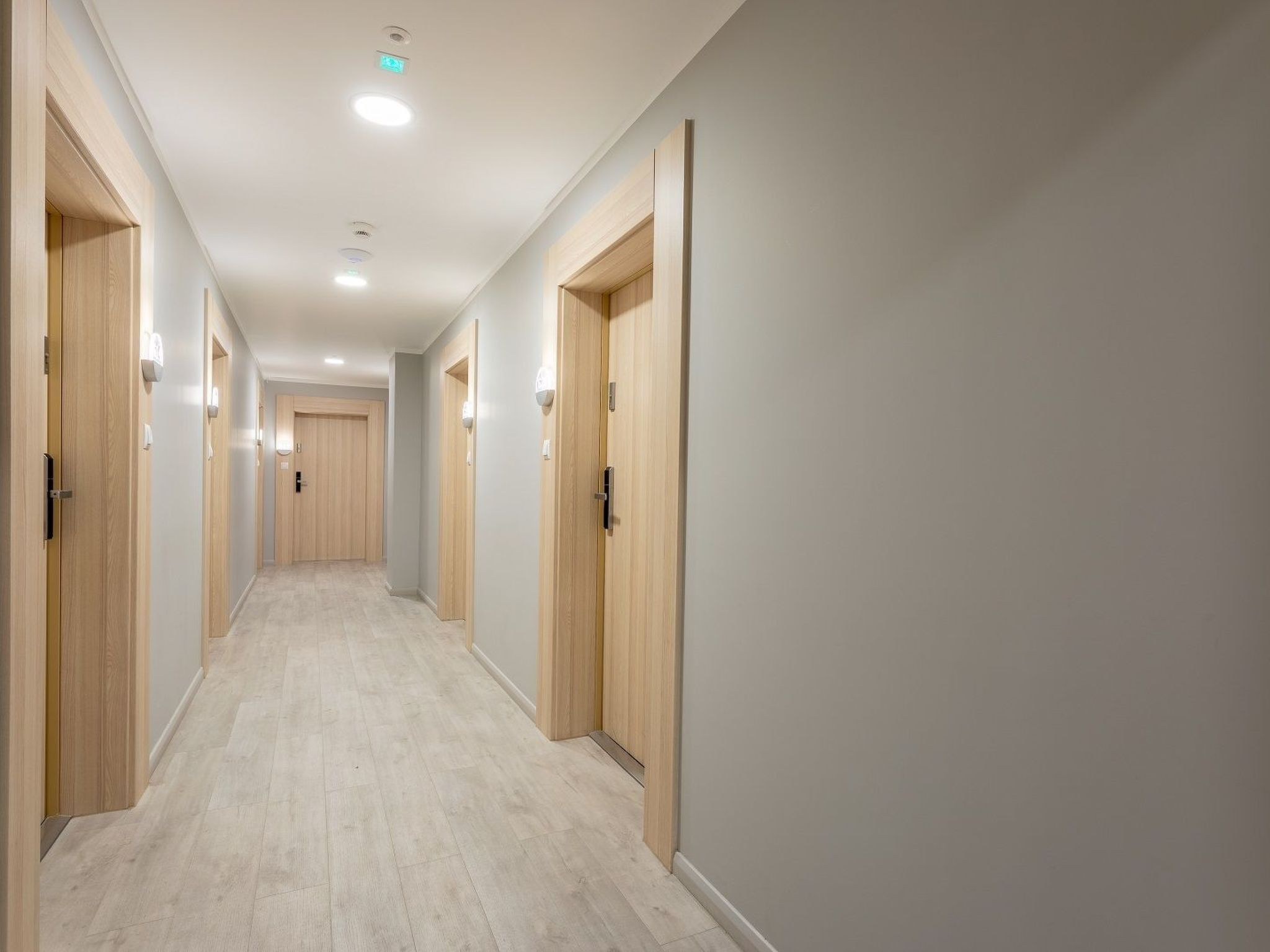 Un pasillo largo y estrecho con puertas de madera a ambos lados, paredes de tono claro y suelo de madera clara.