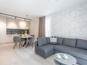 Ein modernes Wohnzimmer mit einem grauen Ecksofa, einem Essbereich mit Tisch und Stühlen sowie einer Küche im Hintergrund.