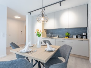 Ett modernt kök med vita skåp, en svart väggbeklädnad bakom arbetsbänken och en matplats med ett träbord där fyra personer kan sitta.