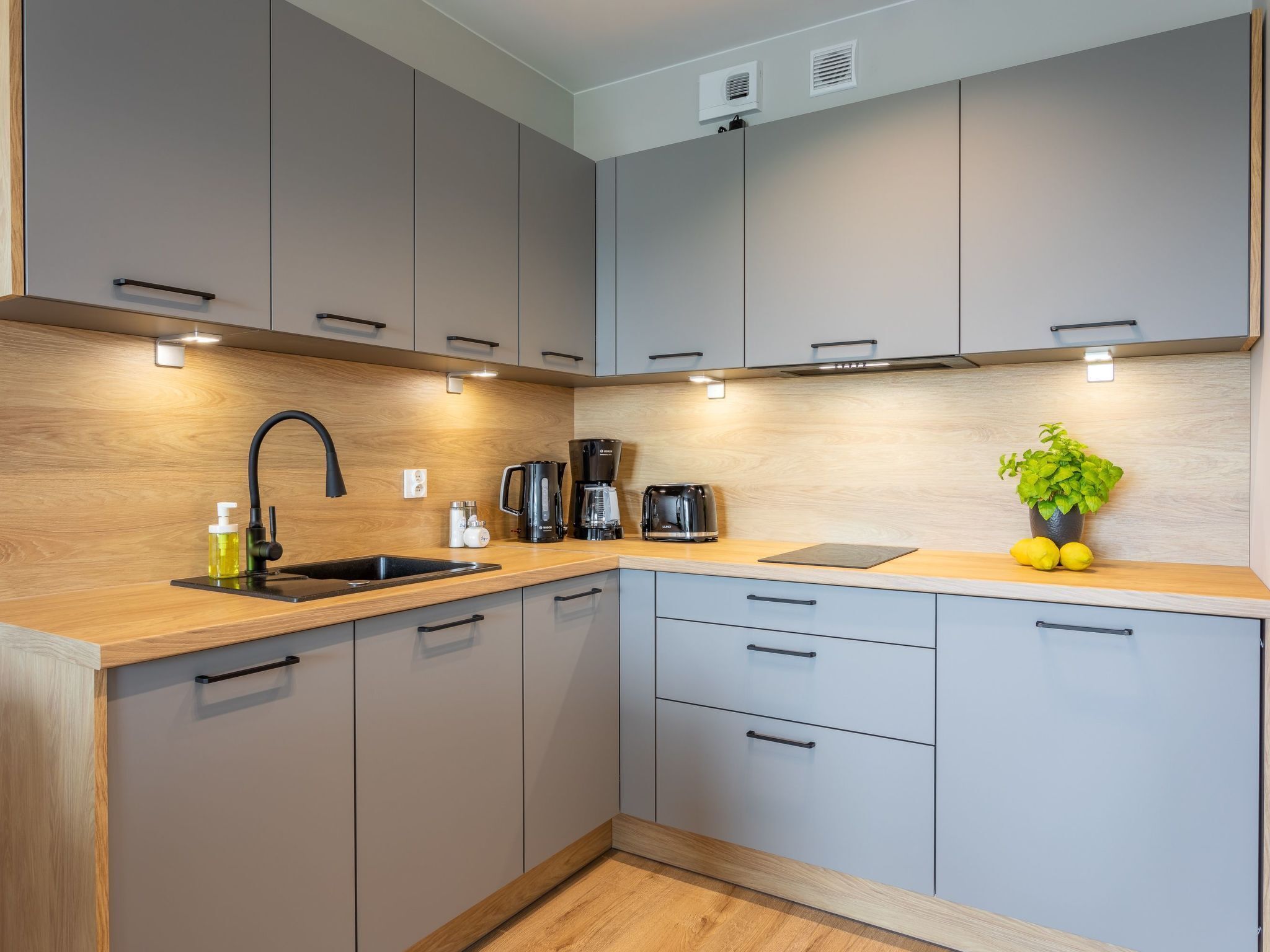 A modern kitchen with grey cabinets, a wooden countertop, a black faucet, a coffee maker, a toaster, and a potted plant with lemons.