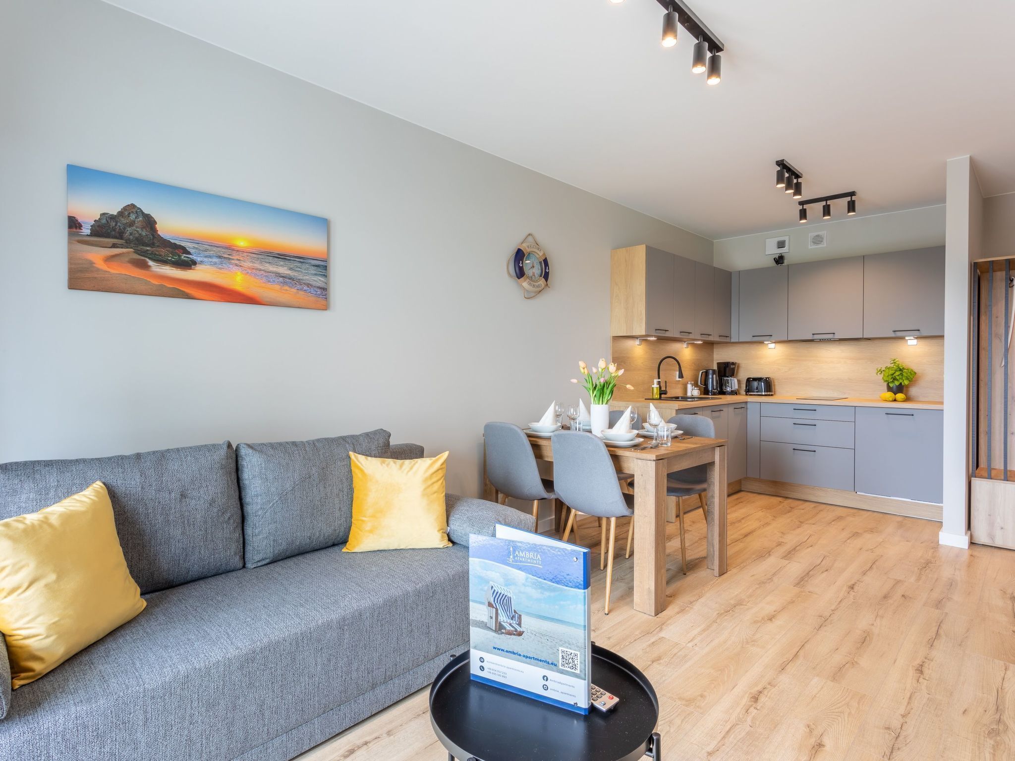 Een moderne woonkamer met een grijze bank, gele kussens, een eettafel voor vier personen en een keuken met grijze kastjes en houten aanrechtbladen.