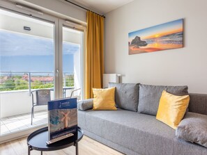 Una sala de estar moderna con un sofá gris, cojines amarillos, un cuadro de un atardecer y vistas de un paisaje urbano a través de un ventanal grande.