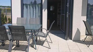 Um pátio externo com mesa e cadeiras, uma porta de correr de vidro e vista da região ao redor.