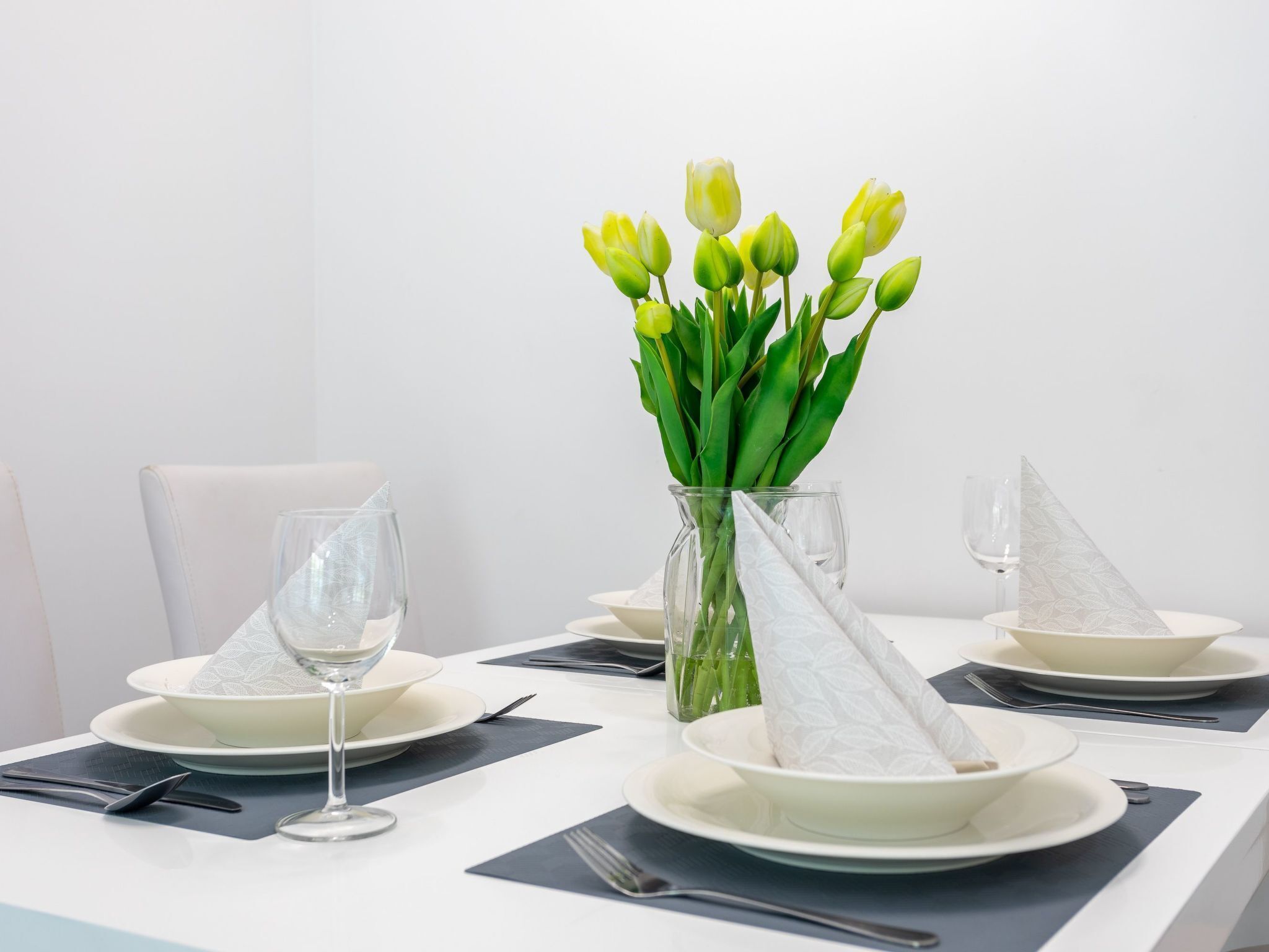 Een eettafel gedekt met witte borden, bestek en een glazen vaas met groene tulpen.