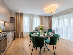A modern dining area with a round table set for four, surrounded by green chairs. A vase with white flowers is on the table. The room has a large window with sheer curtains, a kitchen area to the left, and a sofa with cushions in the background. - mit Balkon an der Ostsee by Interhome (Swinoujscie)