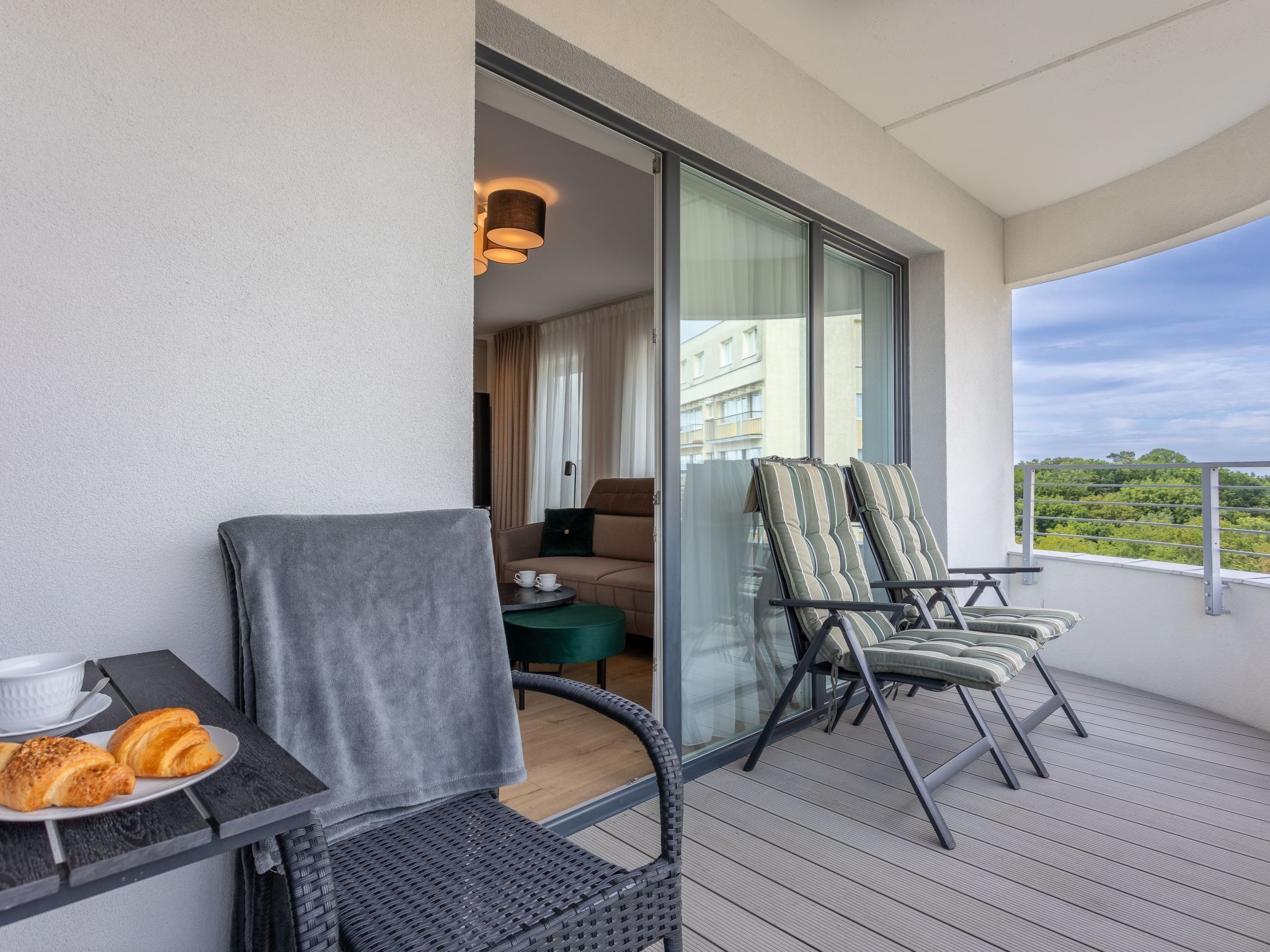 Un balcón con sillas de mimbre, una mesa con croissants y vistas a un paisaje urbano.