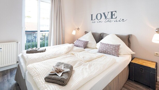 Una cama doble con un almohadón en forma de estrella, dos mesitas de noche, una pared con un letrero que dice 'LOVE is in the air' y una ventana con cortinas.