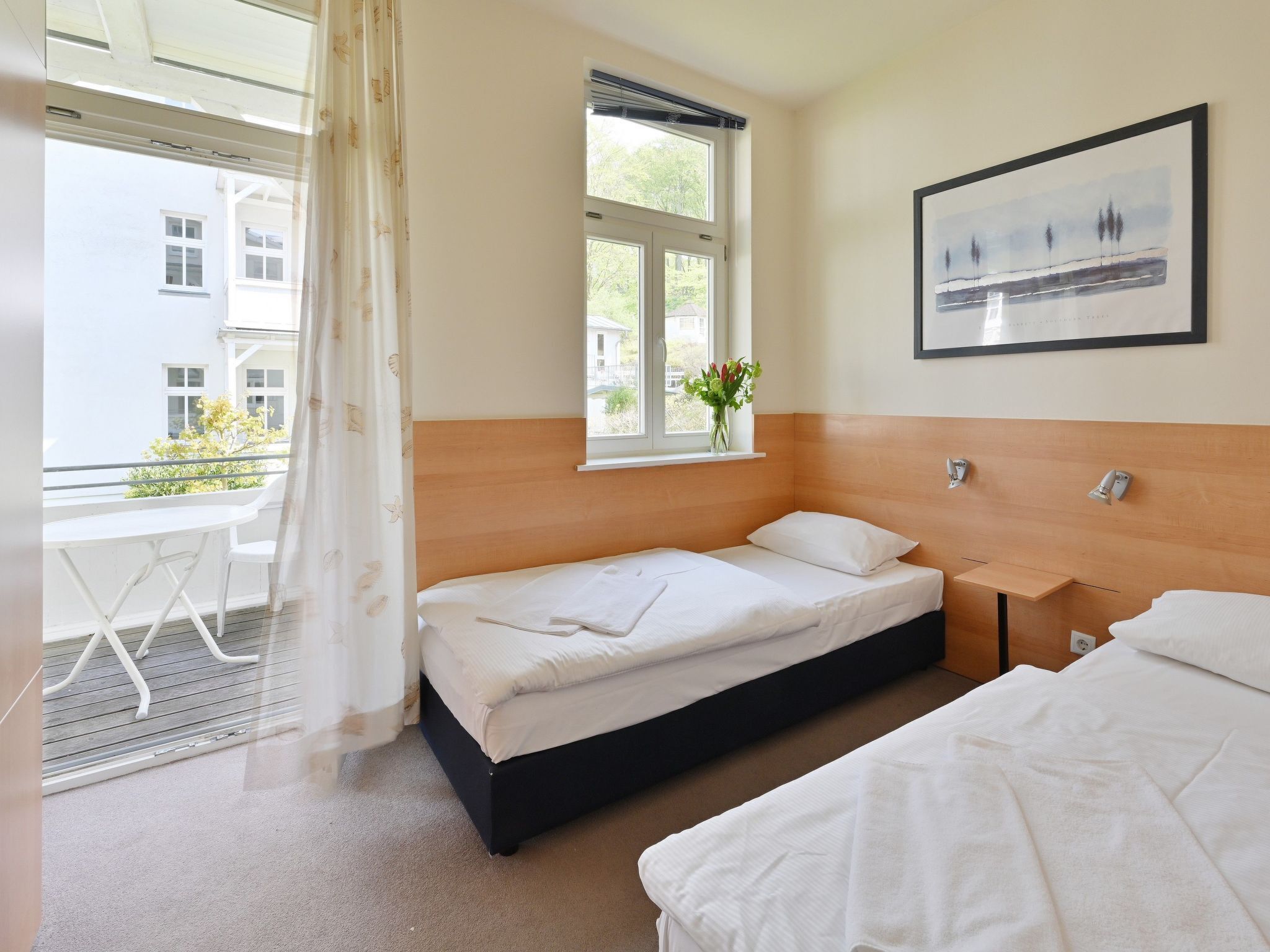 A hotel room with two beds, a window with a view of buildings, a framed picture on the wall, and a small table with a chair.
