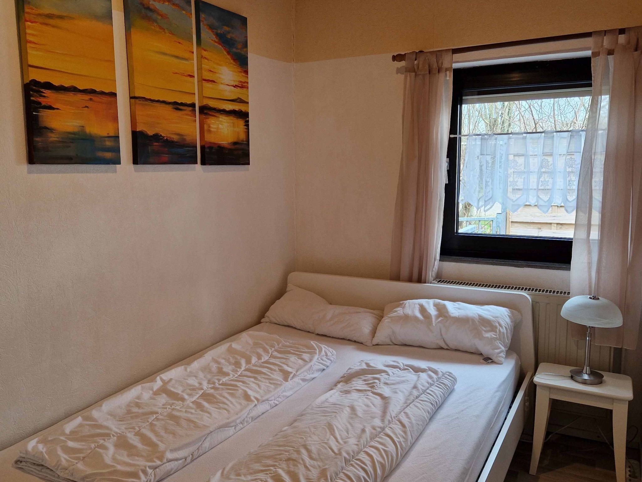 Un lit simple avec un matelas et des oreillers blancs, une table de chevet avec une lampe et une fenêtre avec des rideaux.