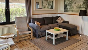 Ett vardagsrum med en grå soffa, en trästol, ett vitt soffbord och ett stort fönster med gardiner.