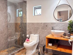 Un baño moderno con ducha de vidrio, inodoro con toalla y flor, tocador de madera con lavamanos y espejo.