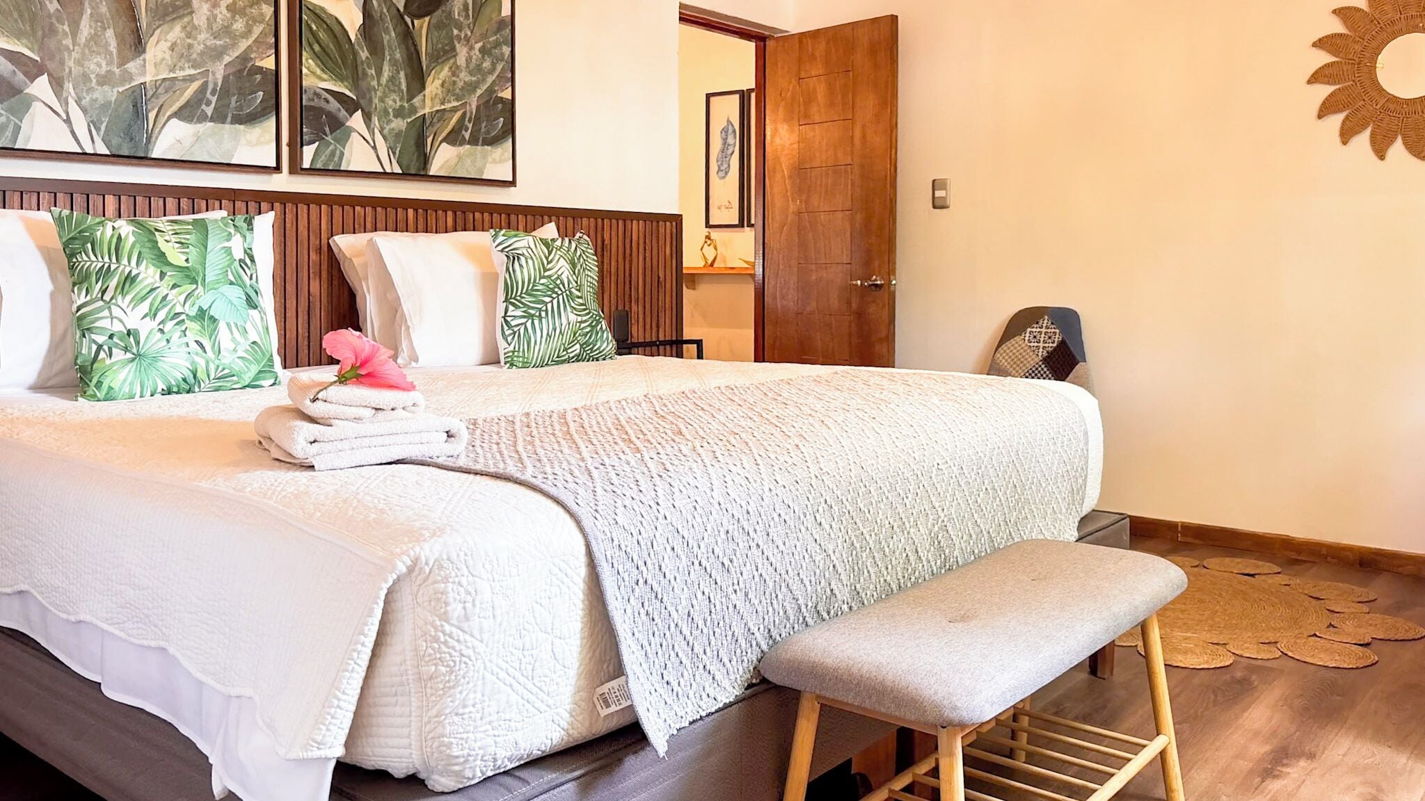 A neatly made bed with a white comforter and pillows, a bench at the foot of the bed, and a wooden door in the background.