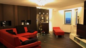 A modern living room with a red sofa, a glass coffee table, and a red ottoman. There is a TV on a stand and a window with a view. - Designerwohnung in denkmalgeschütztem Wasserturm by Interhome (Güstrow)