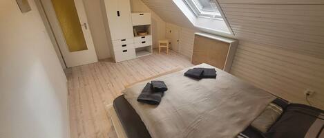A bedroom with a sloped ceiling, a skylight, a bed with a grey blanket, a bedside table, and a wardrobe.