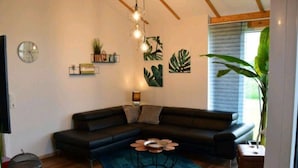 A modern living room with a dark-colored sectional sofa, a round coffee table, and a blue patterned rug. There are wall-mounted shelves with plants and decorative items, and a large potted plant near the window. - Vacation home  by Interhome (Burhave)