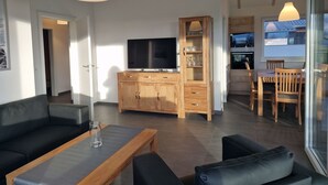 A modern living room with a sofa, coffee table, and TV. There is a wooden cabinet and a dining area with chairs. - G28 Eingezäuntes Ferienhaus mit Infrarotkabine by Interhome (Burhave)