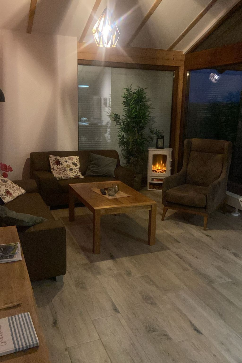A cozy living room with a sofa, armchair, and a wooden coffee table. A fireplace is visible in the background, and a large window offers a view of the outdoors.