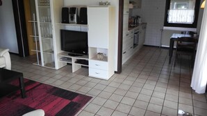En kompakt opholdsstue med fjernsyn, hvide skabe, et spiseområde og et køkken.