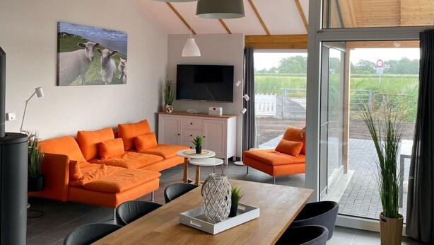 Een moderne woonkamer met een grote oranje bank, een houten eettafel en een aan de muur bevestigde televisie.
