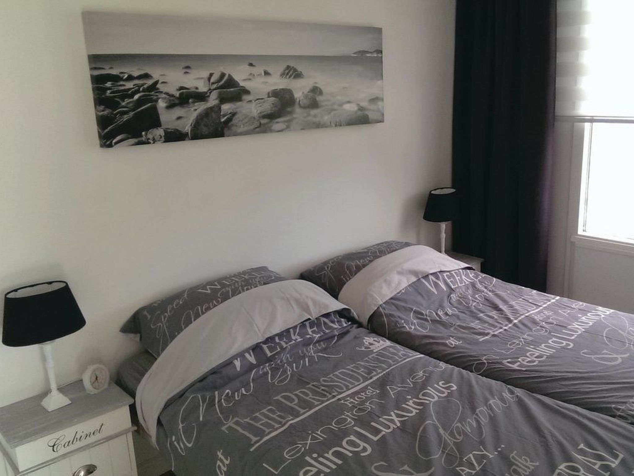 Une chambre à coucher comprenant un lit, une table de chevet, une lampe, une fenêtre avec des rideaux et un tableau accroché au mur.