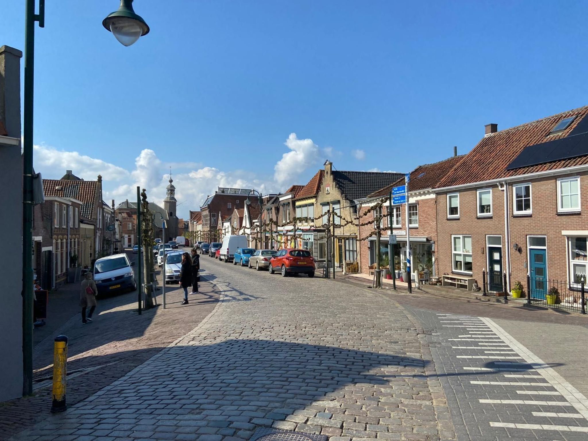 Eine Kopfsteinpflasterstraße gesäumt von traditionellen Gebäuden, einem Kirchturm und geparkten Autos.