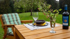 Una mesa de madera con una botella de vino, una copa y un plato con comida, rodeada de follaje y árboles.