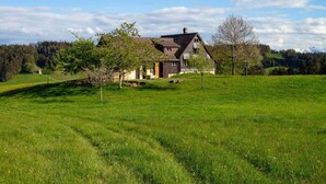 Una casa con techo inclinado, rodeada por un campo verde exuberante y una colina boscosa al fondo.