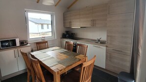 A modern kitchen with wooden cabinets, a dining table with chairs, a microwave, and a window with a view of a building outside. - N4 Haus in Nordseenähe mit Terrasse und Garten by Interhome (Burhave)