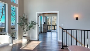 A spacious hallway with wooden flooring, large windows, a potted plant, and a staircase with a black railing. - Skyline Ridge | Modern New Build with Long Range Blue Ridge Mountain Views in Arden (Arden)