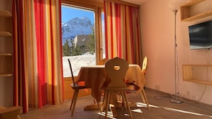A room with a table and chairs, a TV mounted on the wall, and a view of snow-capped mountains through a window with striped curtains. - Apparthotel Pöstli Nr. 106 by Interhome (Maloja)